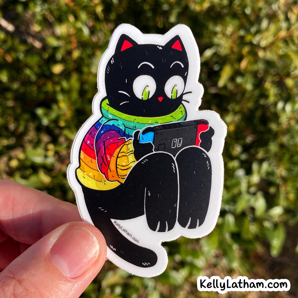 A die-cut sticker featuring a cartoon black cat wearing a rainbow-striped sweater and green scarf, holding a red and blue game controller. The sticker is being held by a hand against a blurred green outdoor background.