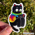 A die-cut sticker featuring a cartoon black cat wearing a rainbow-striped sweater and green scarf, holding a red and blue game controller. The sticker is being held by a hand against a blurred green outdoor background.