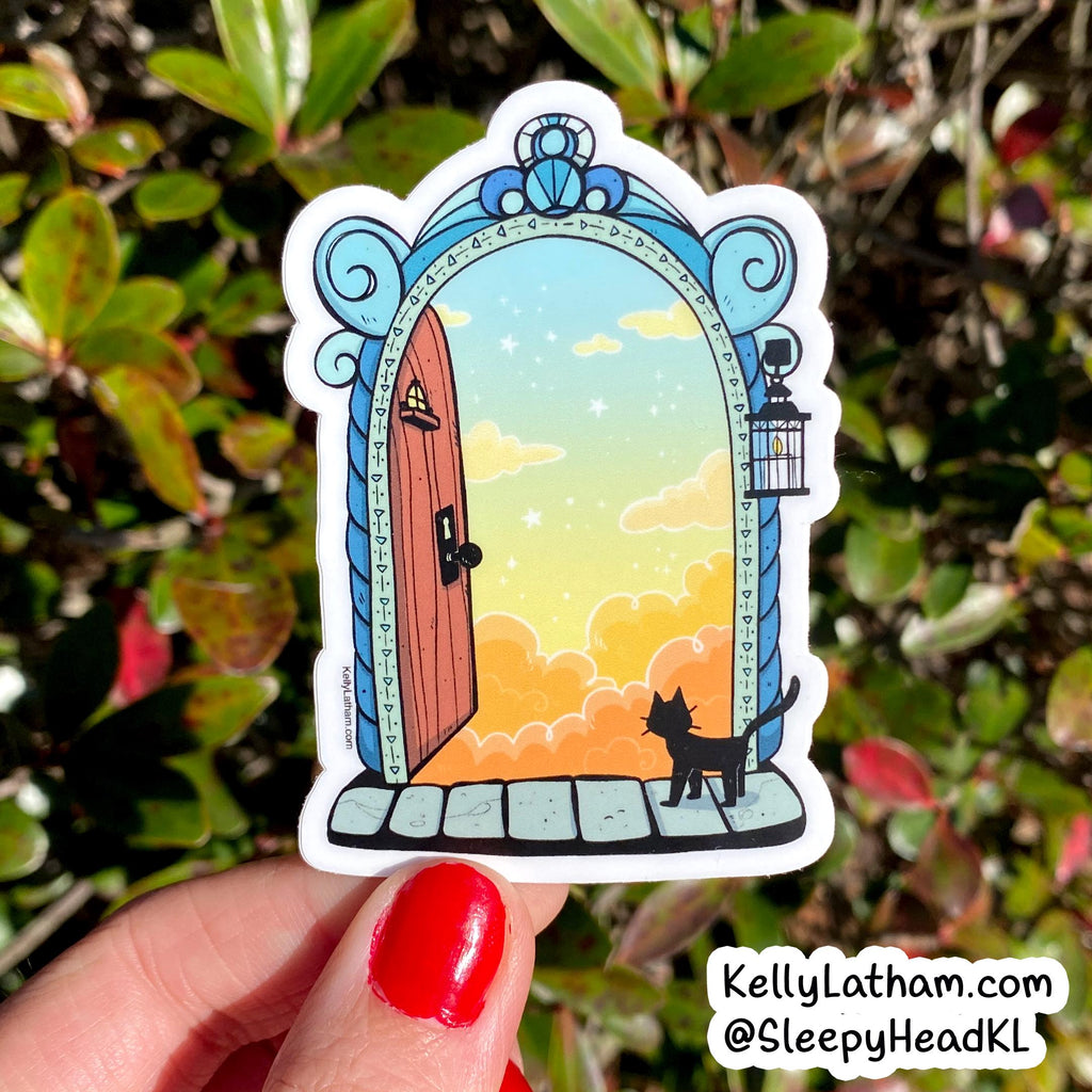 A die-cut sticker held by a hand with red nail polish, featuring an illustration of a black cat looking out of a blue arched doorway at a sunset sky. The background is a blurry garden with green and red leaves.