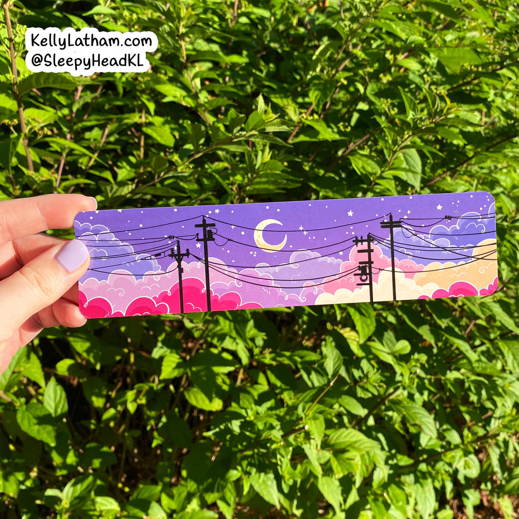 A hand holds a rectangular sticker featuring a whimsical night sky illustration with a crescent moon, stars, clouds, and utility poles against a background of green outdoor foliage.