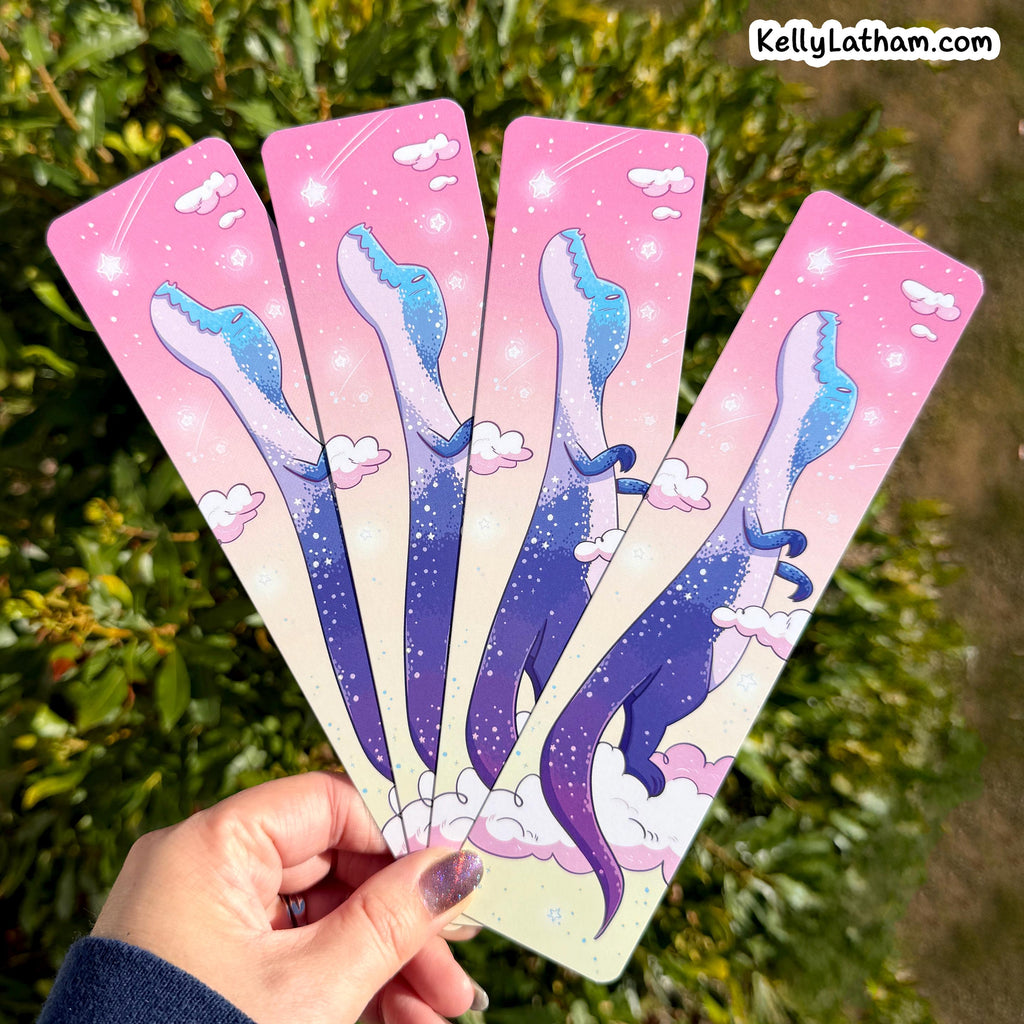 Four matching bookmarks fanned out in a hand, featuring a whimsical illustration of a blue and purple dinosaur climbing among pink clouds on a pink-to-white gradient sky background. The hand is held outdoors in front of green foliage, with a 'KellyLatham.