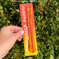 A hand holds a rectangular art print featuring a cartoon bacon strip with the text 'emergency bacon' on a yellow-to-orange gradient background, set against a real green hedge.