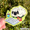 A hand holds a die-cut sticker featuring a cartoon sheep reading in a notebook next to a drink and a red snack bag, with a blurred green outdoor background.