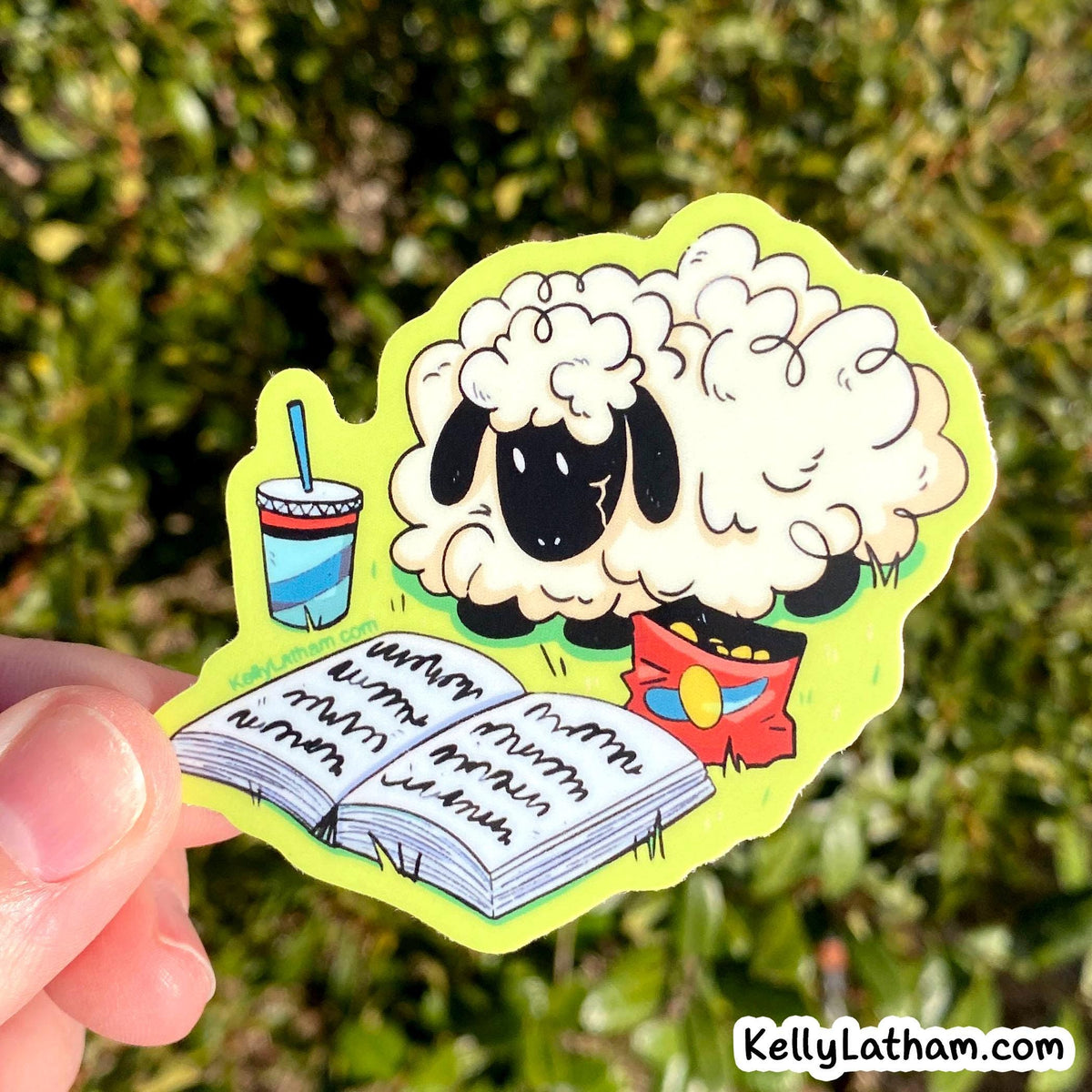 A hand holds a die-cut sticker featuring a cartoon sheep reading in a notebook next to a drink and a red snack bag, with a blurred green outdoor background.