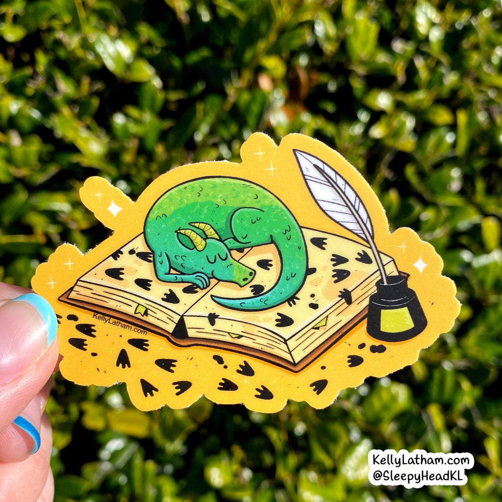 A hand holds a die-cut sticker featuring a sleeping green dragon curled up on an open book with a quill and ink bottle, set against a blurred green outdoor background.