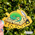 A hand holds a die-cut sticker featuring a sleeping green dragon curled up on an open book with a quill and ink bottle, set against a blurred green outdoor background.