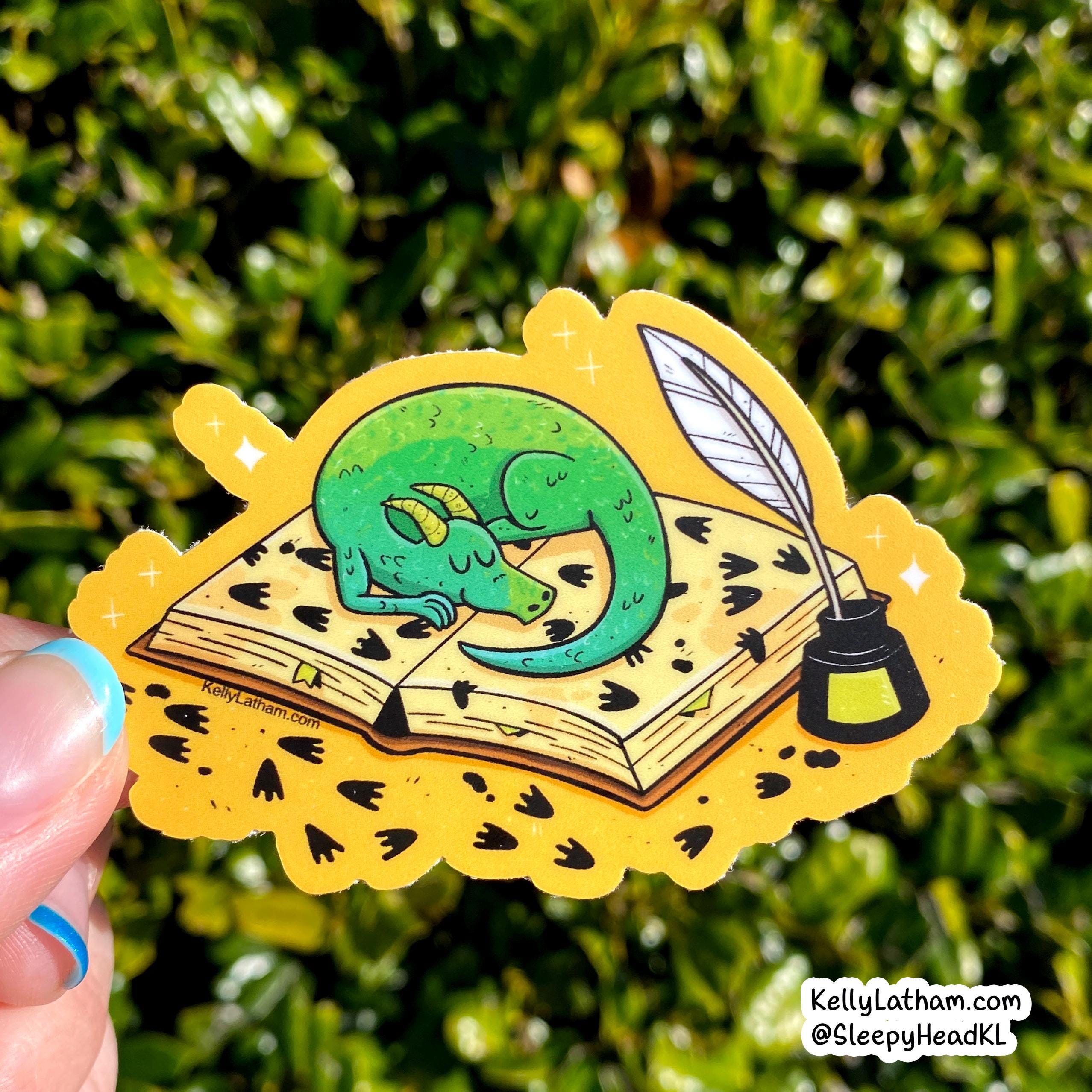 A hand holds a die-cut sticker featuring a sleeping green dragon curled up on an open book with a quill and ink bottle, set against a blurred green outdoor background.