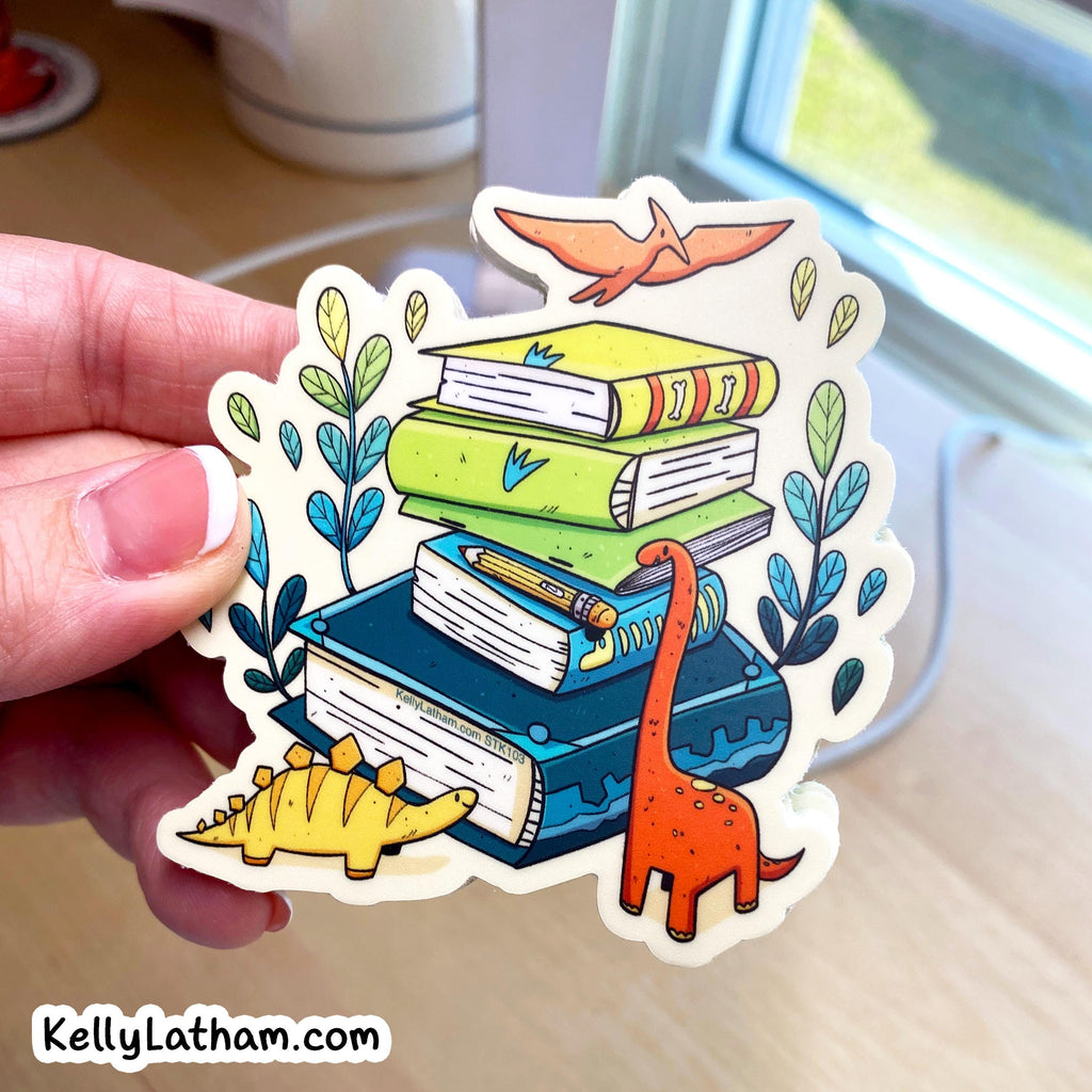A hand holds a die-cut sticker featuring an illustration of stacked books with dinosaurs and plants. The sticker has a white border and is shown against a blurred indoor background near a window.