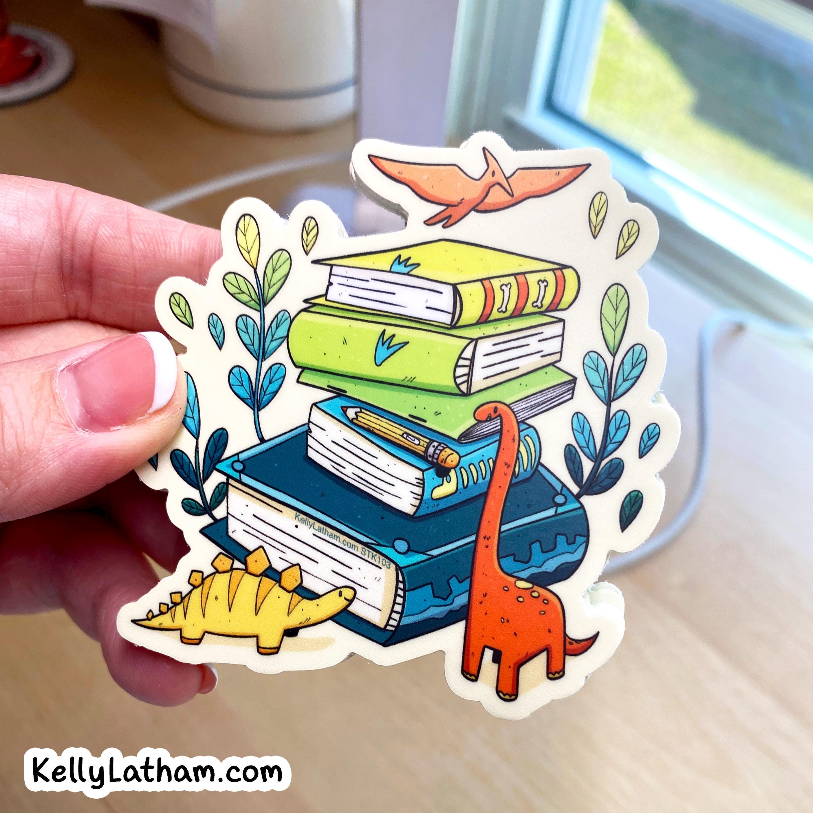 A hand holds a die-cut sticker featuring an illustration of stacked books with dinosaurs and plants. The sticker has a white border and is shown against a blurred indoor background near a window.