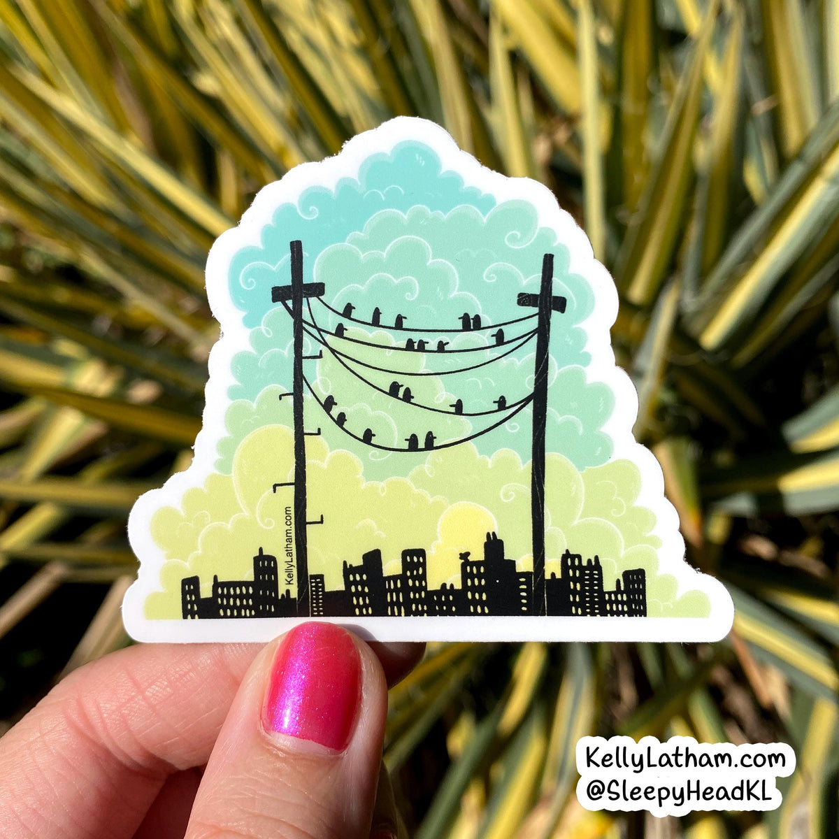 A hand with pink glitter nail polish holds a die-cut sticker featuring a stylized city skyline under a pastel sky with clouds, power lines, and birds, with green/yellow variegated leaves blurred in the background.