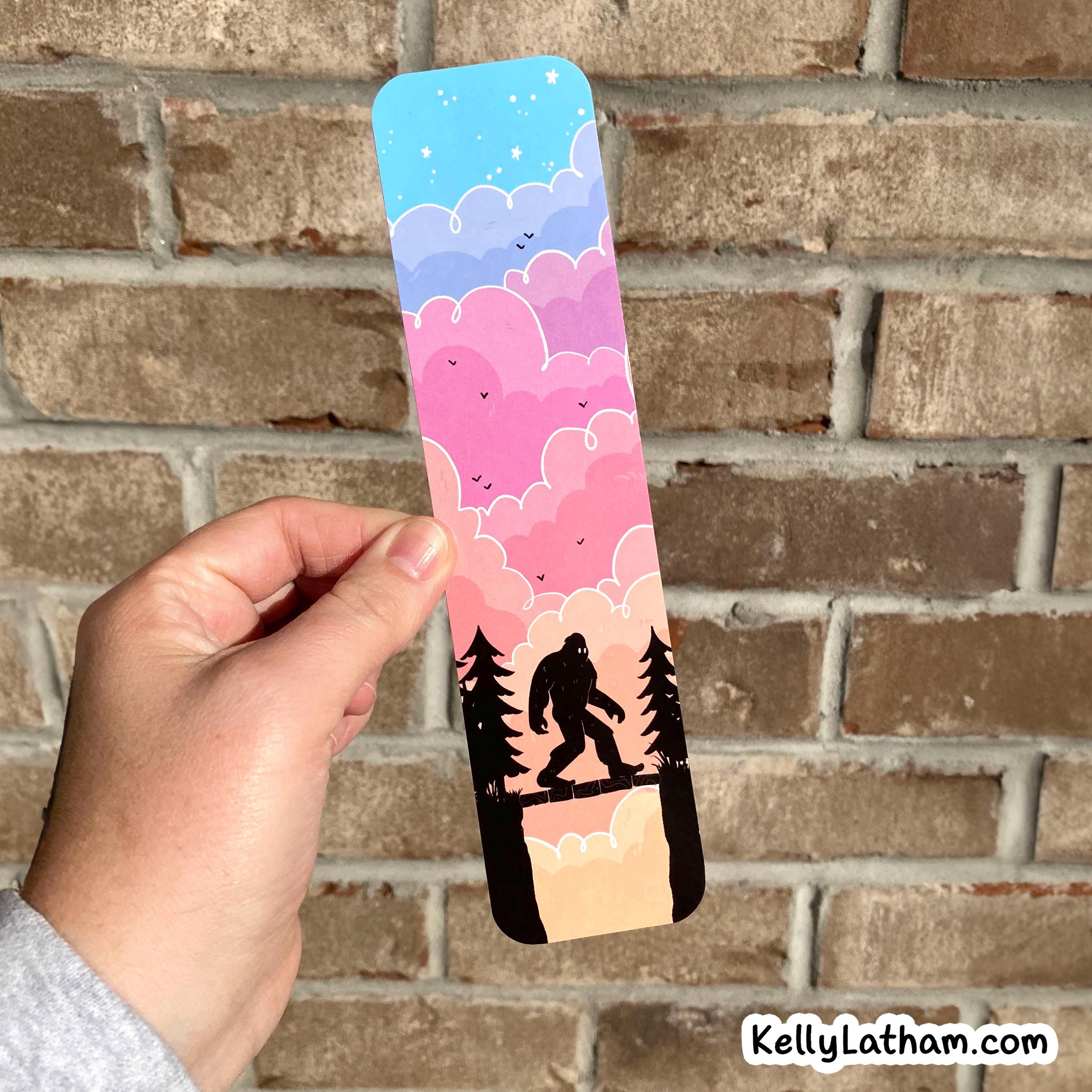 A hand holds a slim rectangular printed card or bookmark in front of a brick wall. The printed design shows a pastel sky with blue, pink, and lavender clouds, small birds, tree silhouettes, and a black silhouette figure walking across a cliff.
