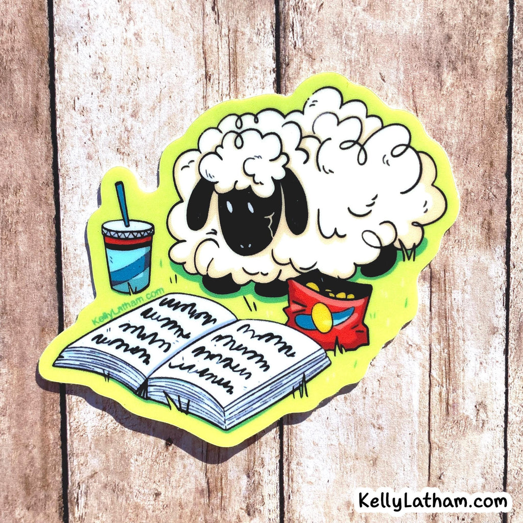 A die-cut sticker featuring a cartoon sheep with curly white wool, a black face, and a red bowl, shown with an open book and a striped drink. The sticker is photographed on a rustic wood plank background.