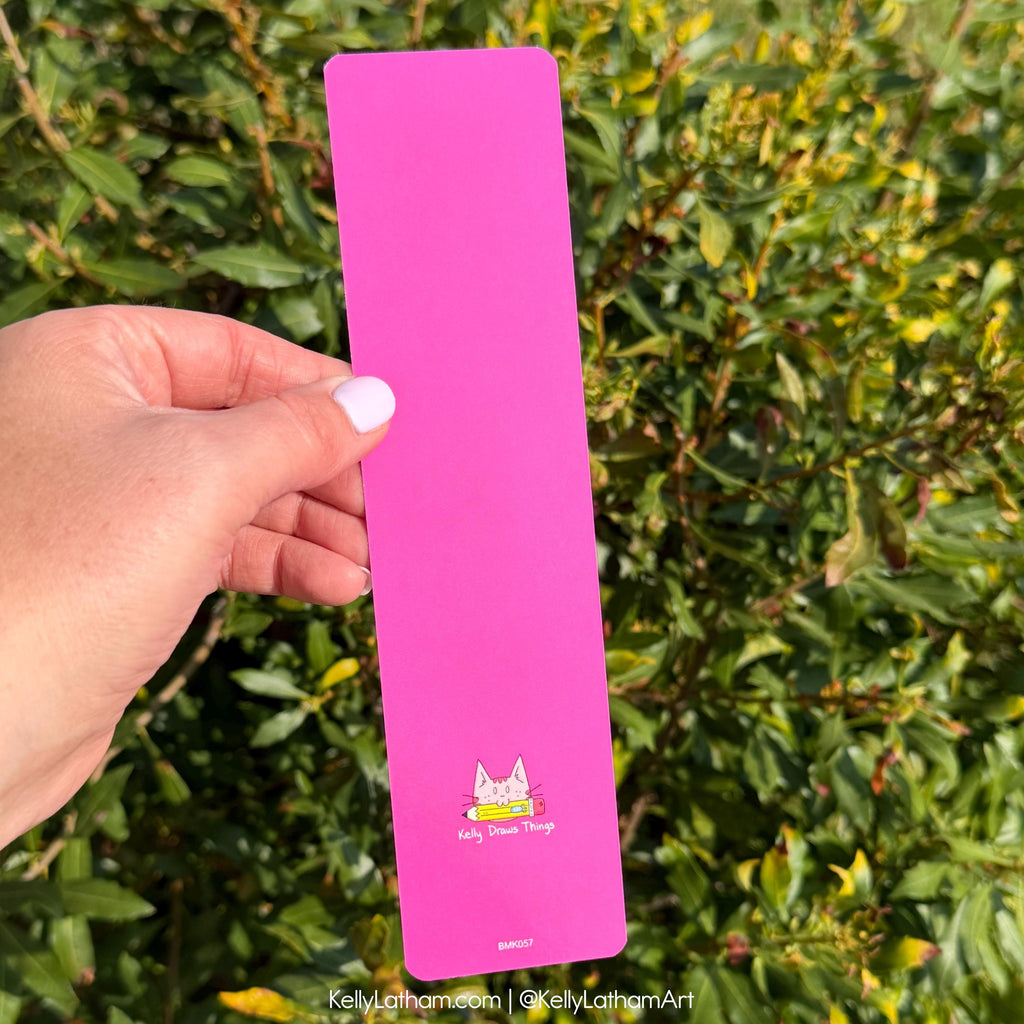 A hand holds a bright pink rectangular metal bookmark or sign outdoors in front of green foliage. The pink item has a small cat illustration and the text 'Kelly Latham Things' near the bottom.