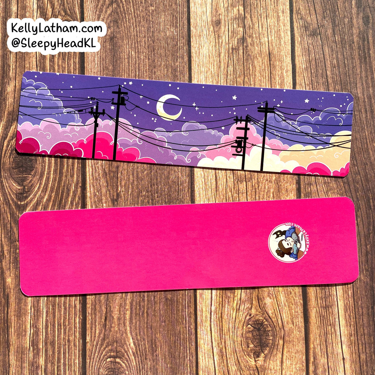 Two rectangular printed items laid flat on a wood surface. The top item has a pastel night-sky illustration with clouds, stars, a crescent moon, and black utility poles; the bottom item is bright pink with a small circular cartoon logo near one end.