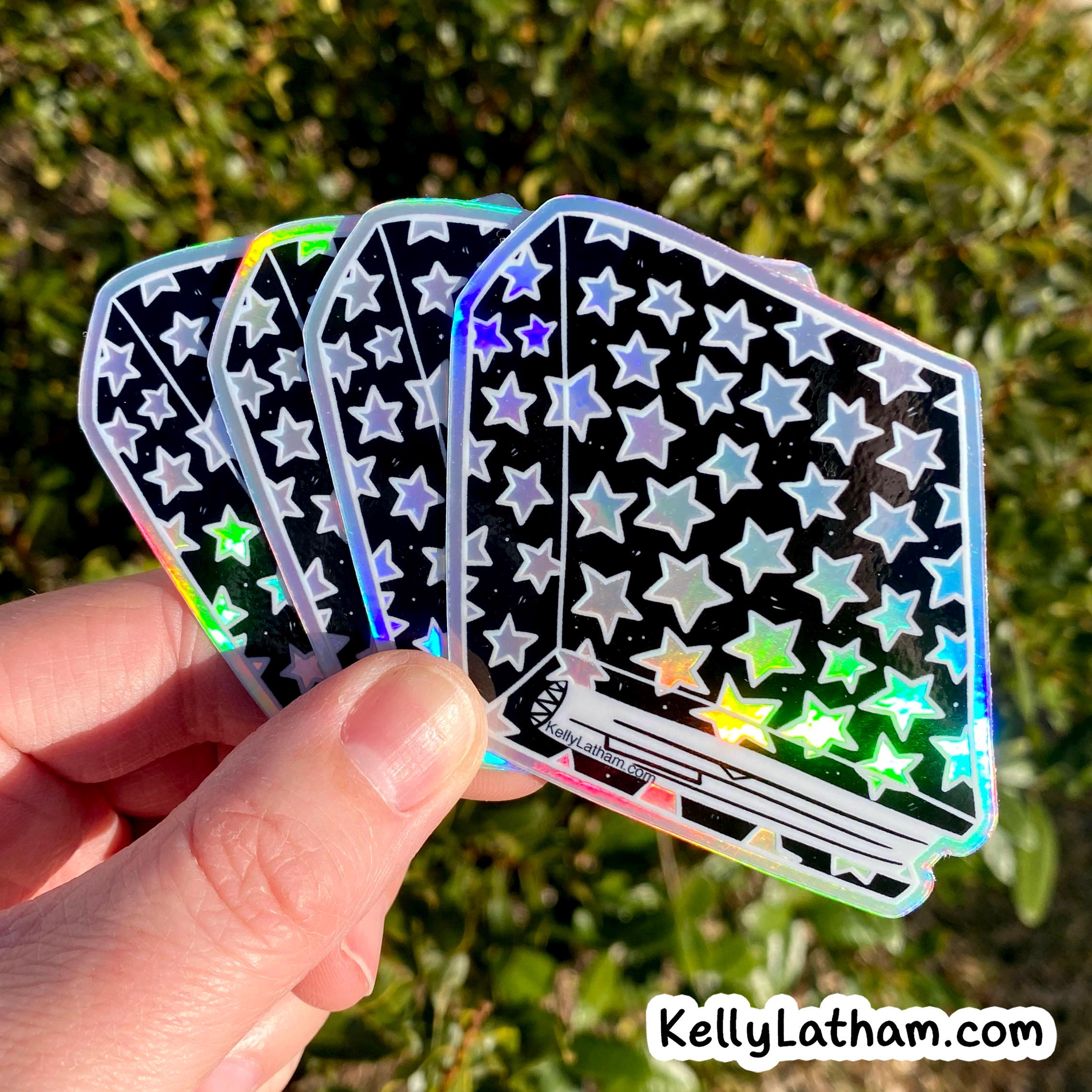 A hand holds four holographic stickers with a black background and white star pattern, photographed outdoors against blurred green foliage. A white 'KellyLatham.