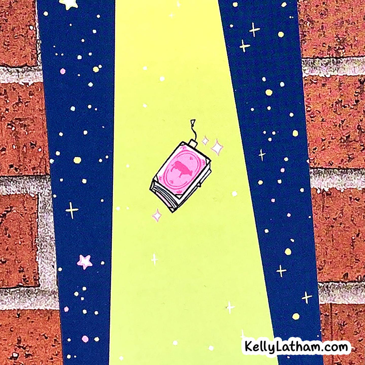 A stylized illustration of a pink book or notebook floating in a neon yellow vertical beam, framed by dark blue starry panels and a red brick wall background. A small 'KellyLatham.