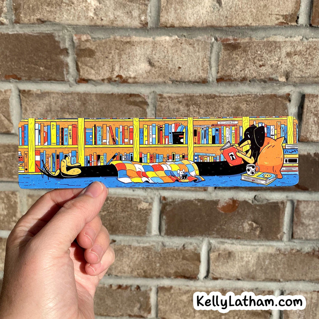 A hand holds a long rectangular illustrated plaque or sign in front of a brick wall. The artwork shows a cozy library scene with bookshelves, books, a person reading, and a dog lying under a checkered blanket.