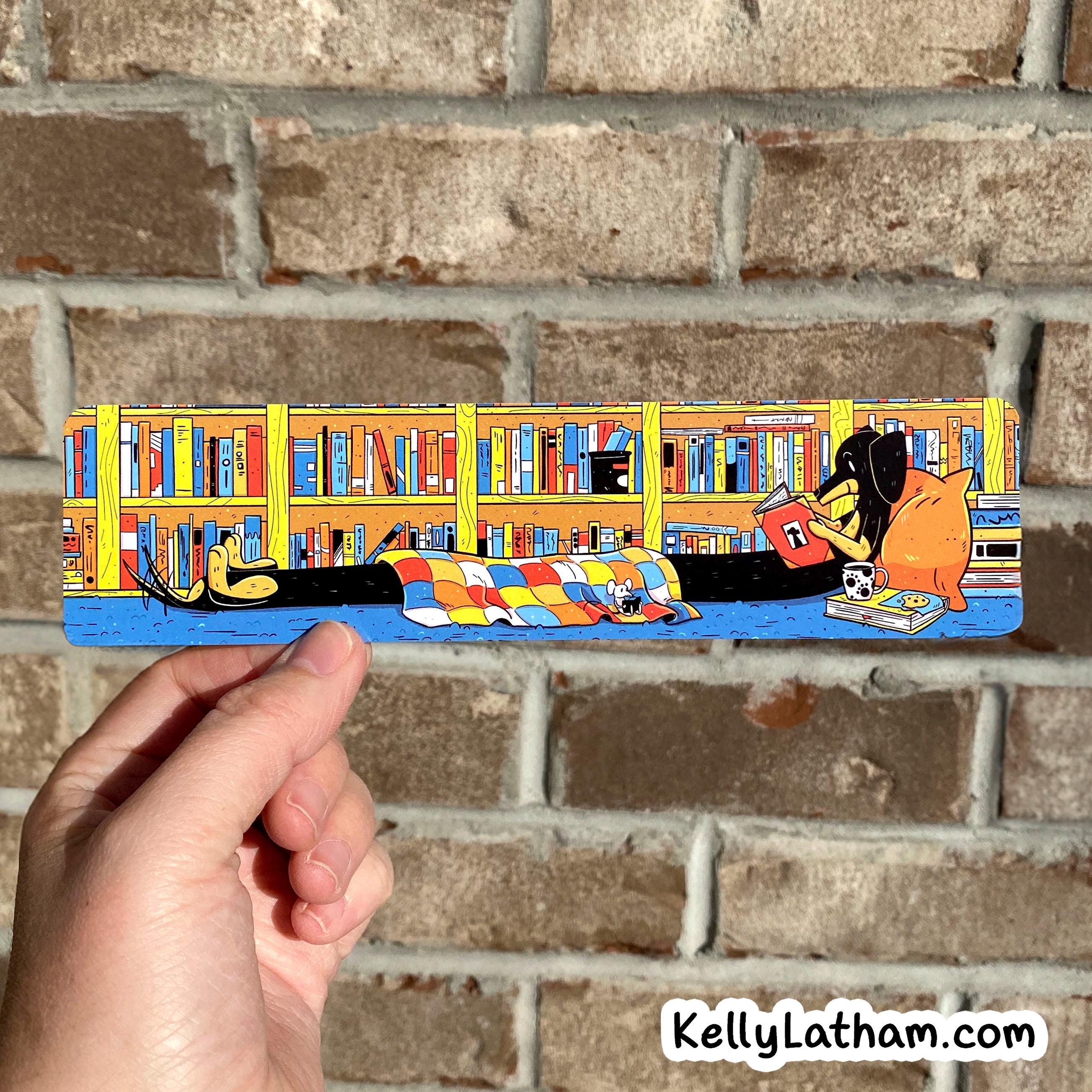 A hand holds a long rectangular illustrated plaque or sign in front of a brick wall. The artwork shows a cozy library scene with bookshelves, books, a person reading, and a dog lying under a checkered blanket.