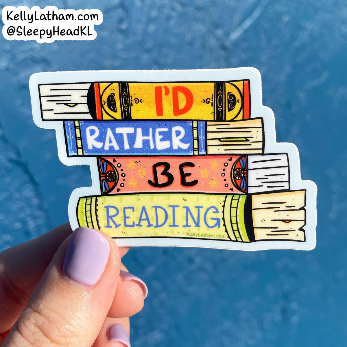 A hand with lavender nail polish holds a die-cut sticker shaped like stacked books with the words “I'D RATHER BE READING” in colorful lettering, against a bright blue textured background. A store watermark is visible in the top left corner.