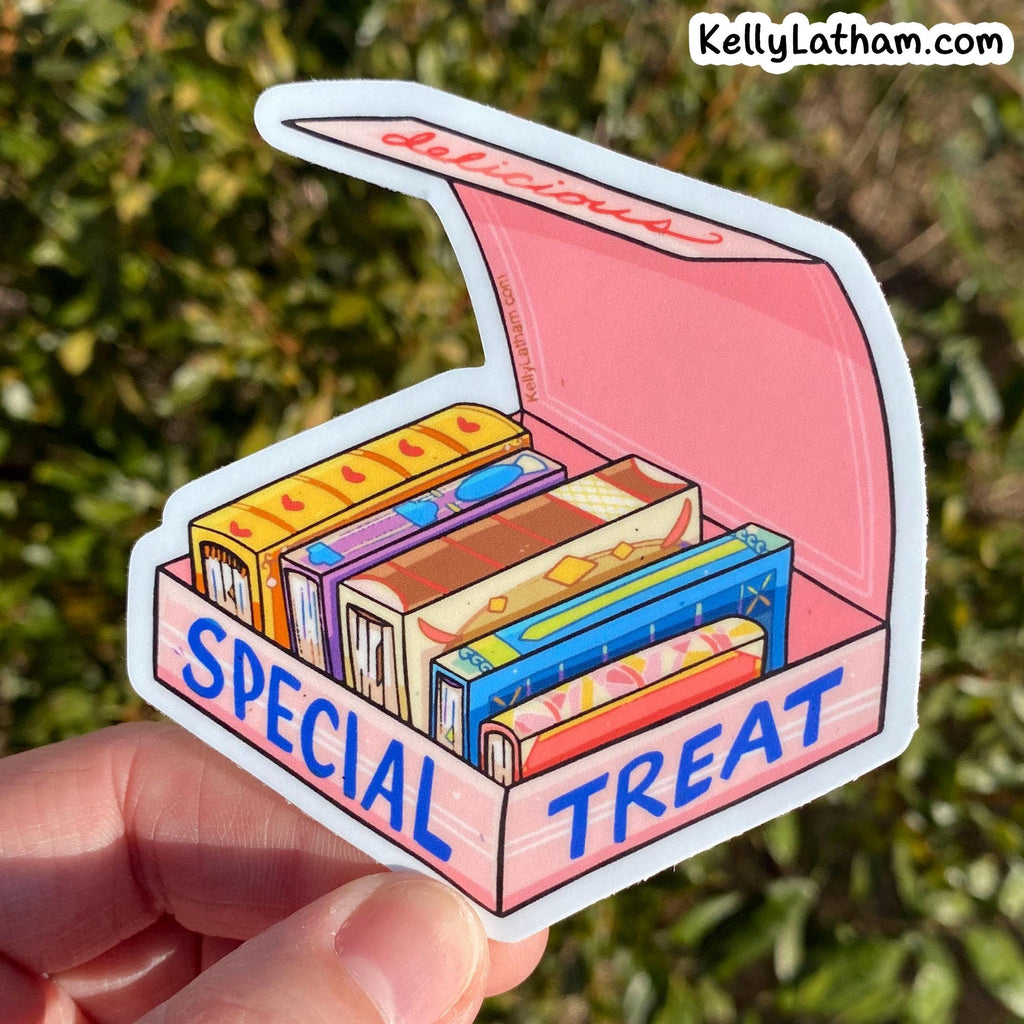 A hand holds a die-cut sticker shaped like an open pink treat box filled with colorful illustrated candy bars. The sticker says 'SPECIAL TREAT' in blue letters, and the background is an out-of-focus outdoor green hedge.