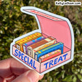 A hand holds a die-cut sticker shaped like an open pink treat box filled with colorful illustrated candy bars. The sticker says 'SPECIAL TREAT' in blue letters, and the background is an out-of-focus outdoor green hedge.