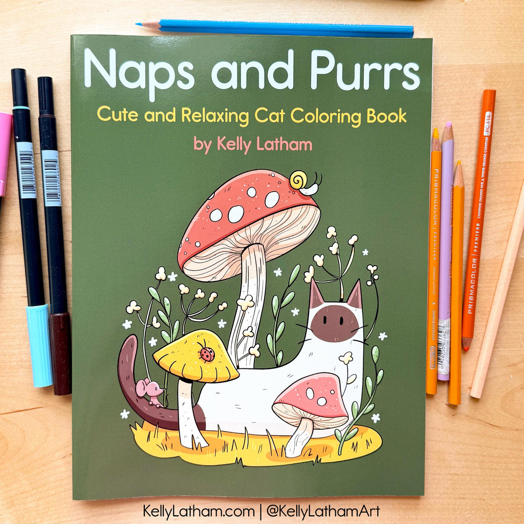 The front cover of the 'Naps and Purrs' coloring book is displayed on a light wood desk, surrounded by drawing tools including markers on the left and colored pencils on the right. The book cover is olive green with a colorful illustration of a white cat, pink and yellow mushrooms, and small flowers.