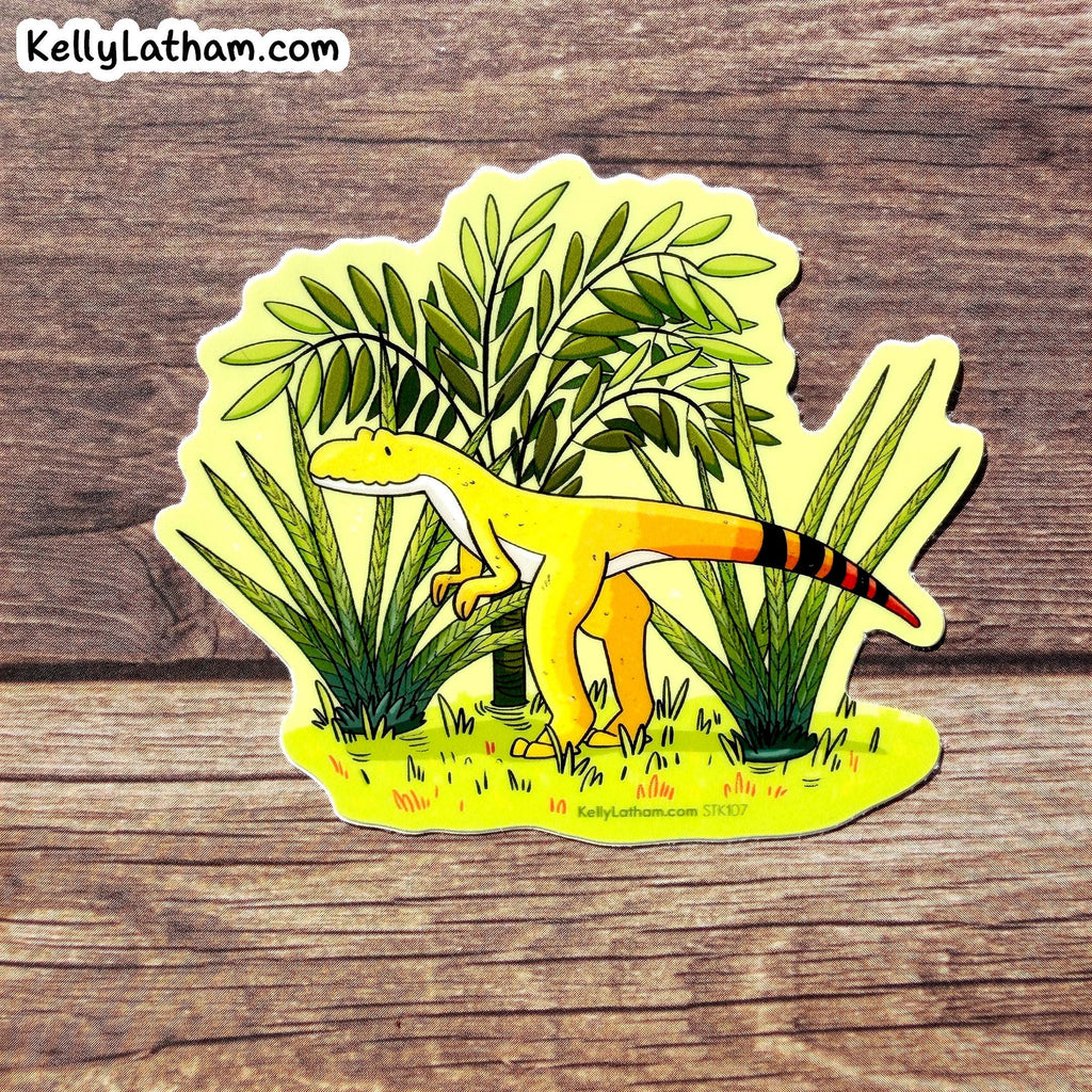 A die-cut sticker featuring a cartoon yellow theropod dinosaur among green plants, placed on a rustic brown wood surface. The sticker has the brand name KellyLatham.