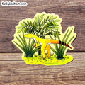 A die-cut sticker featuring a cartoon yellow theropod dinosaur among green plants, placed on a rustic brown wood surface. The sticker has the brand name KellyLatham.