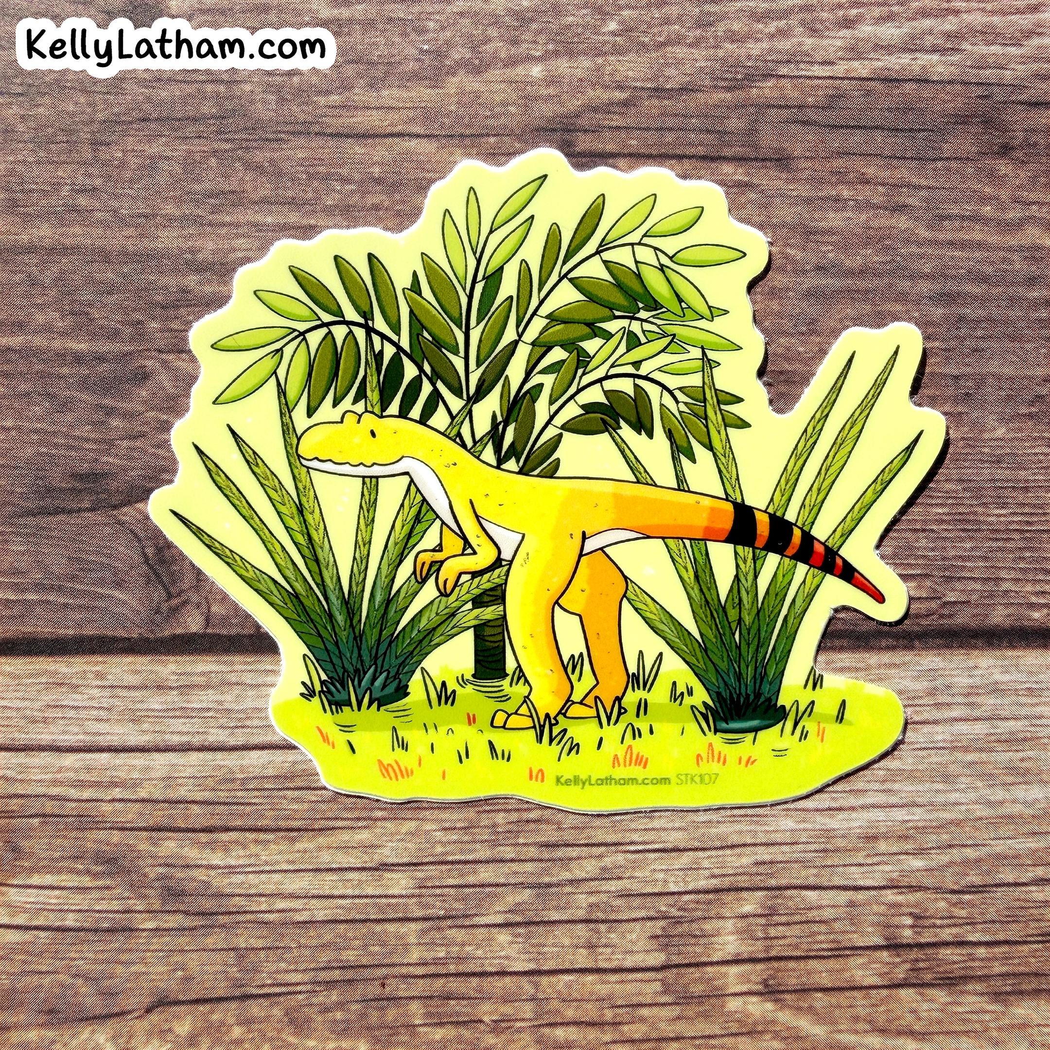 A die-cut sticker featuring a cartoon yellow theropod dinosaur among green plants, placed on a rustic brown wood surface. The sticker has the brand name KellyLatham.