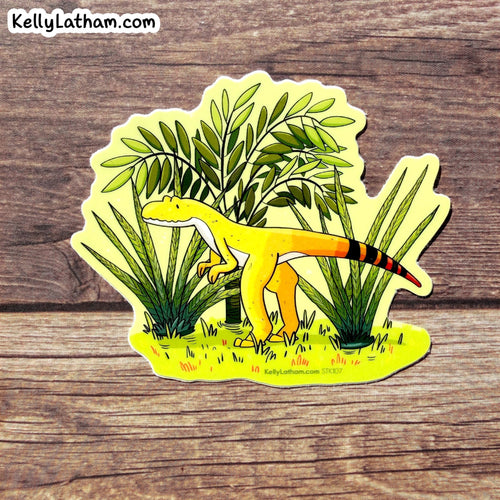 A die-cut sticker featuring a cartoon yellow theropod dinosaur among green plants, placed on a rustic brown wood surface. The sticker has the brand name KellyLatham.