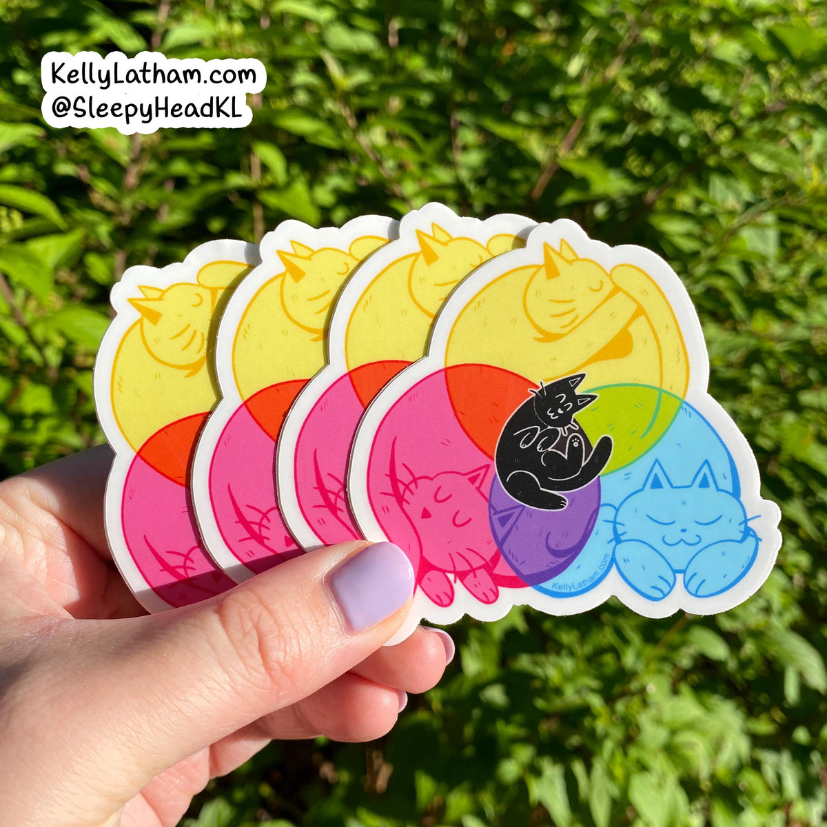 A hand holds a fan of five die-cut stickers featuring cartoon cats curled up inside pastel-colored circles. The stickers show different color combinations: yellow, orange, red, pink, purple, green, blue, and black.