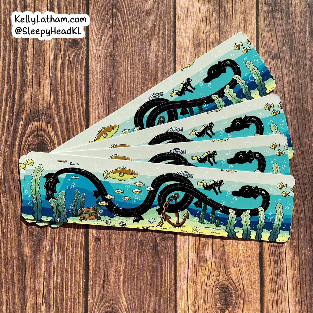 Four rectangular coasters with an illustrated underwater scene featuring a black serpent, divers, fish, kelp, and an anchor, arranged in a fan on a brown wood plank background. A white label in the top-left corner reads KellyLatham.