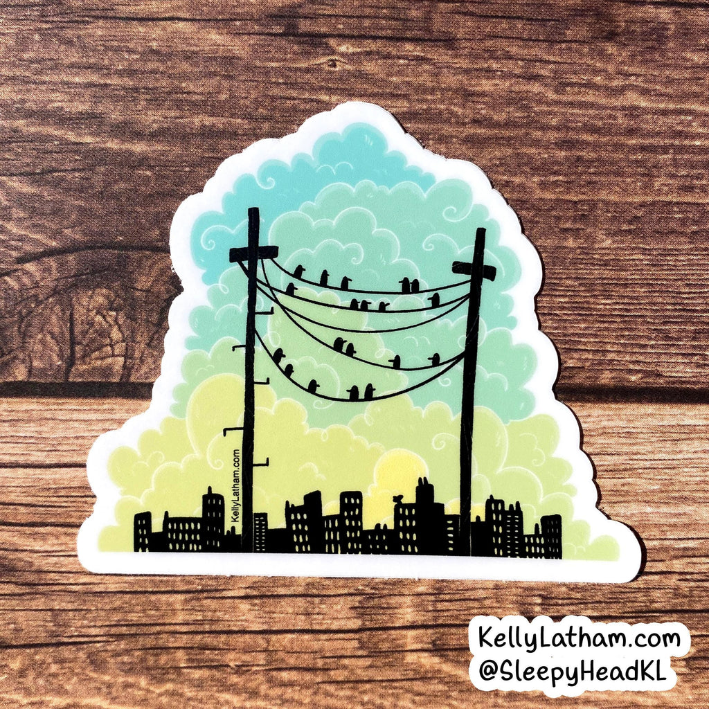 A die-cut sticker featuring a stylized city skyline with black silhouettes, two utility poles, and birds perched on wires against a pastel sky gradient from teal to yellow, shown on a brown wood surface.