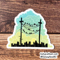A die-cut sticker featuring a stylized city skyline with black silhouettes, two utility poles, and birds perched on wires against a pastel sky gradient from teal to yellow, shown on a brown wood surface.
