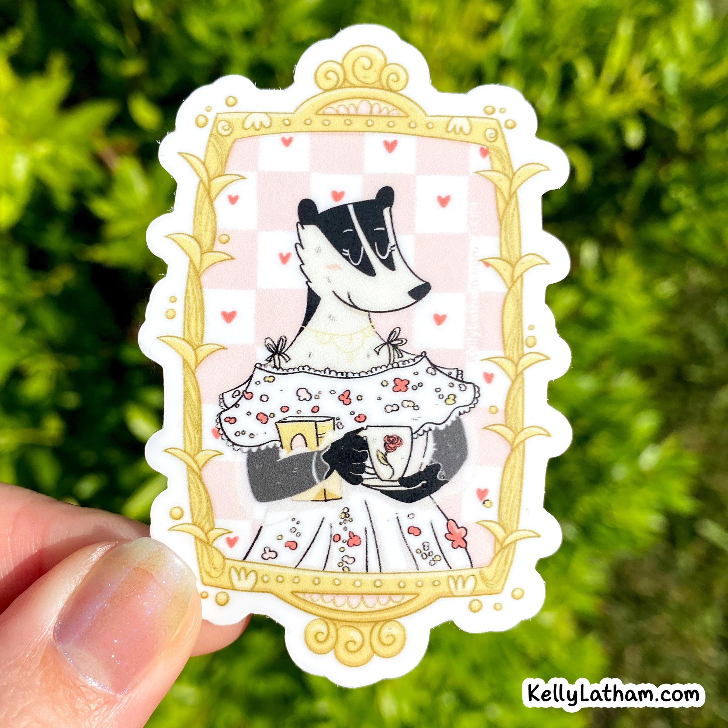 A hand holds a die-cut sticker featuring a stylized badger in a white dress with red hearts, framed by a gold ornate border on a pink checkered background. The scene is photographed outdoors in front of blurred green foliage.