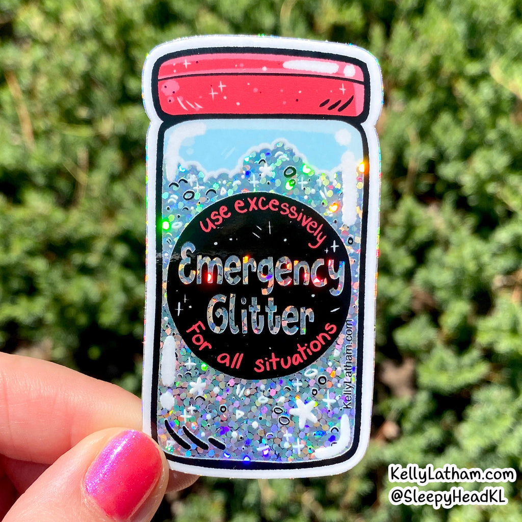 A hand with pink nail polish holds a die-cut sticker shaped like an emergency glitter jar. The sticker features a red lid, a black center circle with the text 'Emergency Glitter For all situations', and a background of blue glitter with multicolor reflective flakes.
