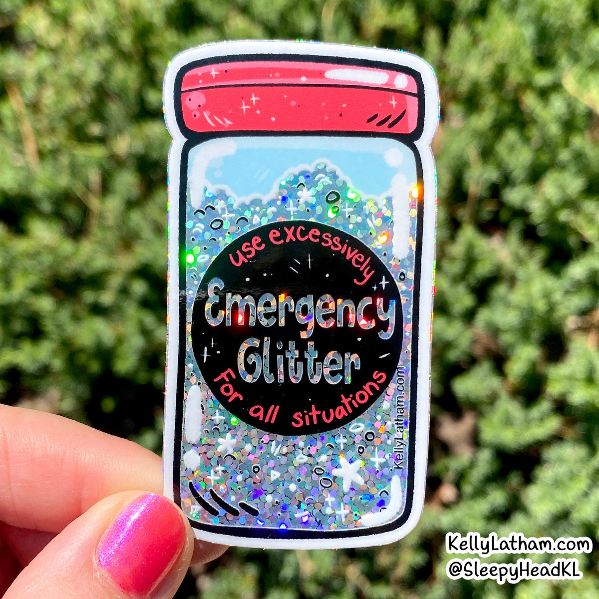 A hand with pink nail polish holds a die-cut sticker shaped like an emergency glitter jar. The sticker features a red lid, a black center circle with the text 'Emergency Glitter For all situations', and a background of blue glitter with multicolor reflective flakes.