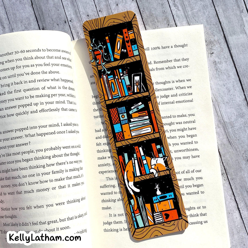 A rectangular wooden bookmark or bookplate featuring a colorful illustration of a cozy library shelf with books, a globe, and a cat, displayed on top of open book pages on a gray wood background.