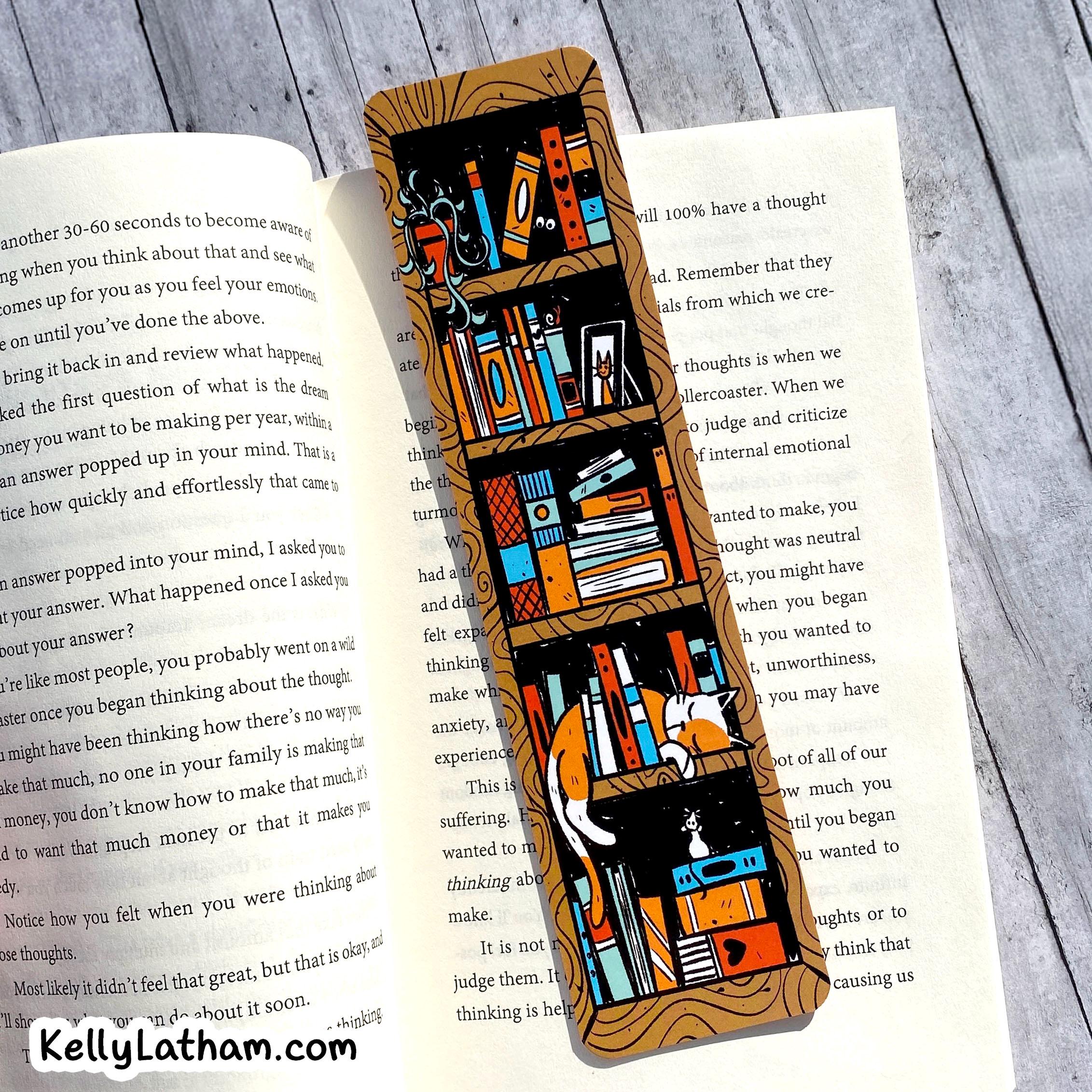 A rectangular wooden bookmark or bookplate featuring a colorful illustration of a cozy library shelf with books, a globe, and a cat, displayed on top of open book pages on a gray wood background.