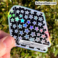 A hand holds a die-cut holographic sticker featuring a black background with a pattern of silver, blue, and pink stars, set against a blurred green outdoor background.