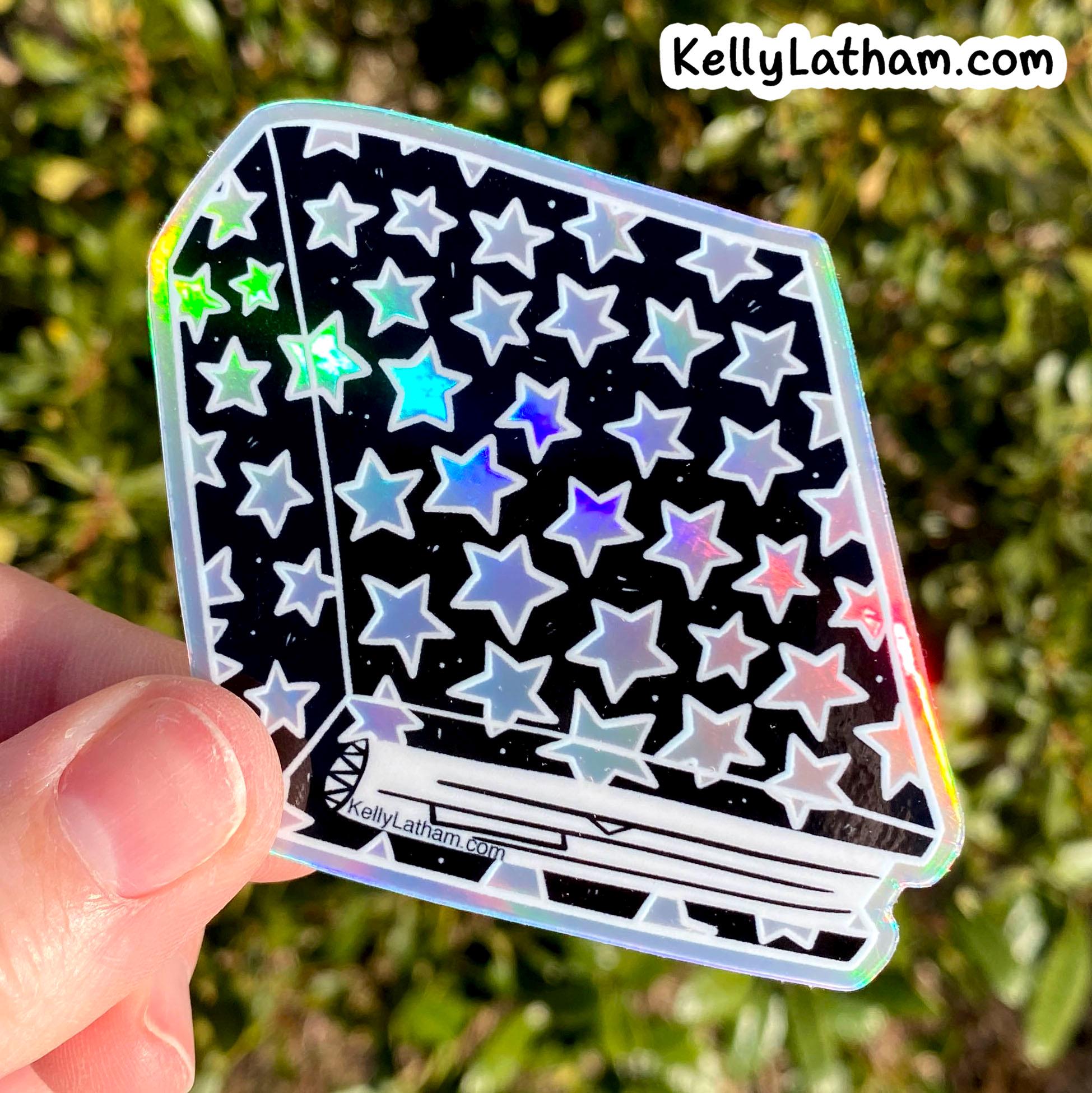 A hand holds a die-cut holographic sticker featuring a black background with a pattern of silver, blue, and pink stars, set against a blurred green outdoor background.