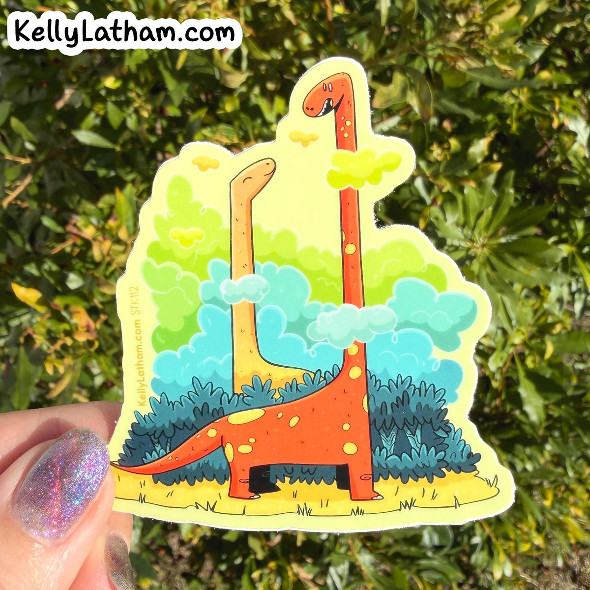 A die-cut sticker featuring two long-necked dinosaurs in a colorful landscape, held by a hand with glittery nail polish against a background of green foliage. The sticker has 'KellyLatham.