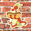 A die-cut sticker featuring a whimsical red dragon carrying a hot dog with mustard and relish, and holding a bag of chips, shown against a red brick wall background. The sticker has a cream border and includes the text 'KellyLatham.