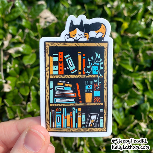 A die-cut sticker featuring a whimsical illustration of a black cat sleeping on a wooden bookshelf filled with colorful books and plants, held by a hand against a background of green outdoor foliage.