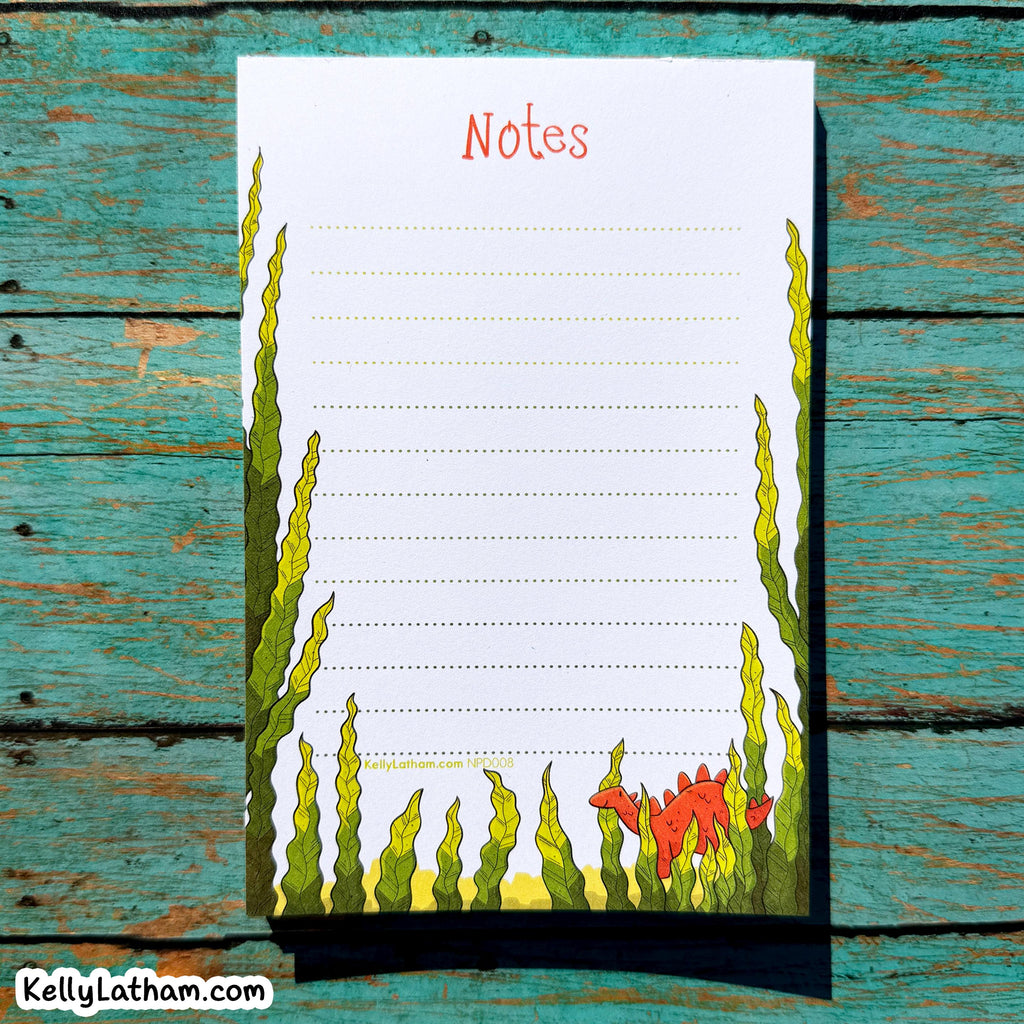 A vertical notepad sheet titled "Notes" with lined writing space, bordered by illustrated green seaweed and a small red fish at the bottom, placed on a distressed turquoise wooden surface.
