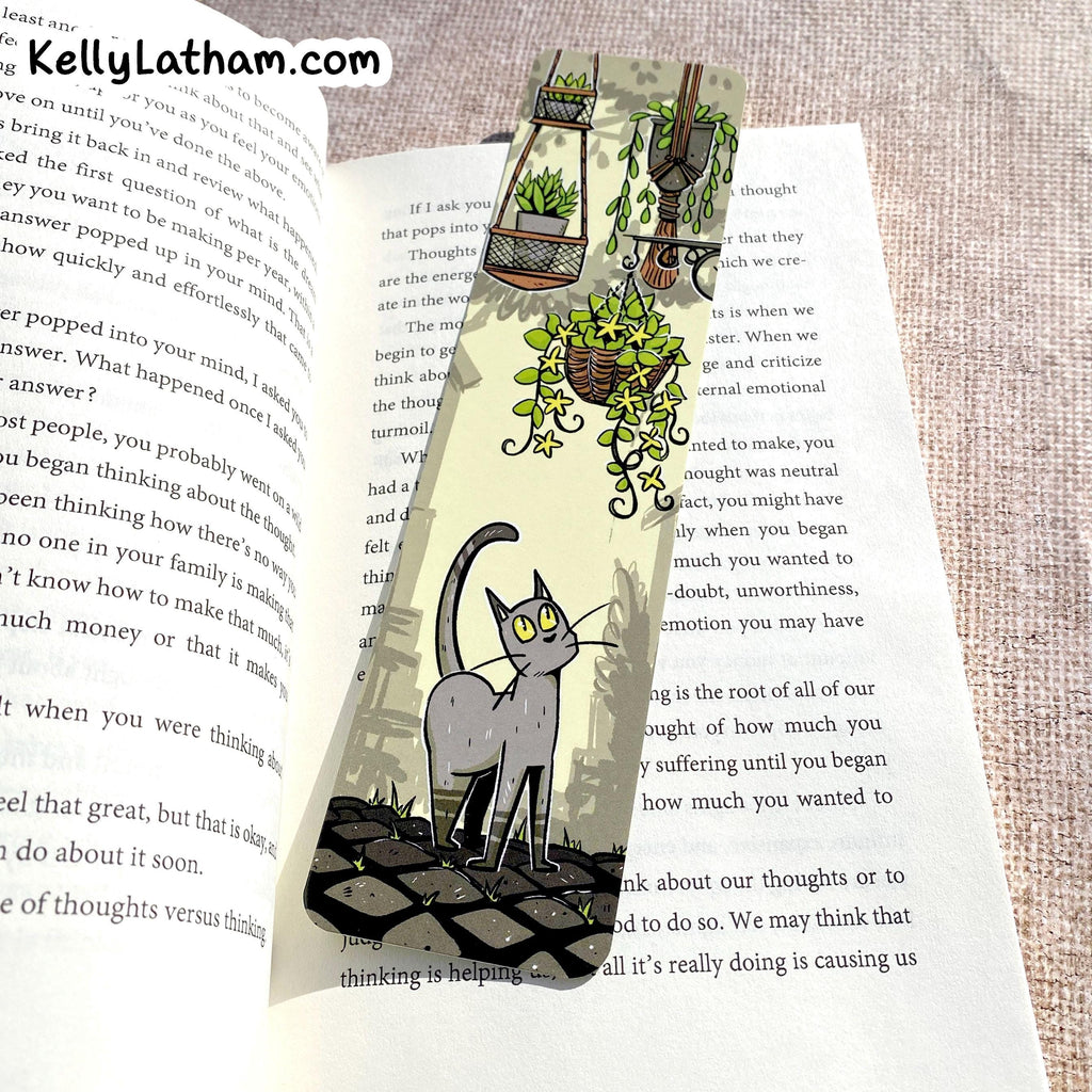 A handmade paper bookmark featuring a black and white cat and potted plants, displayed on an open book over a textured linen background.