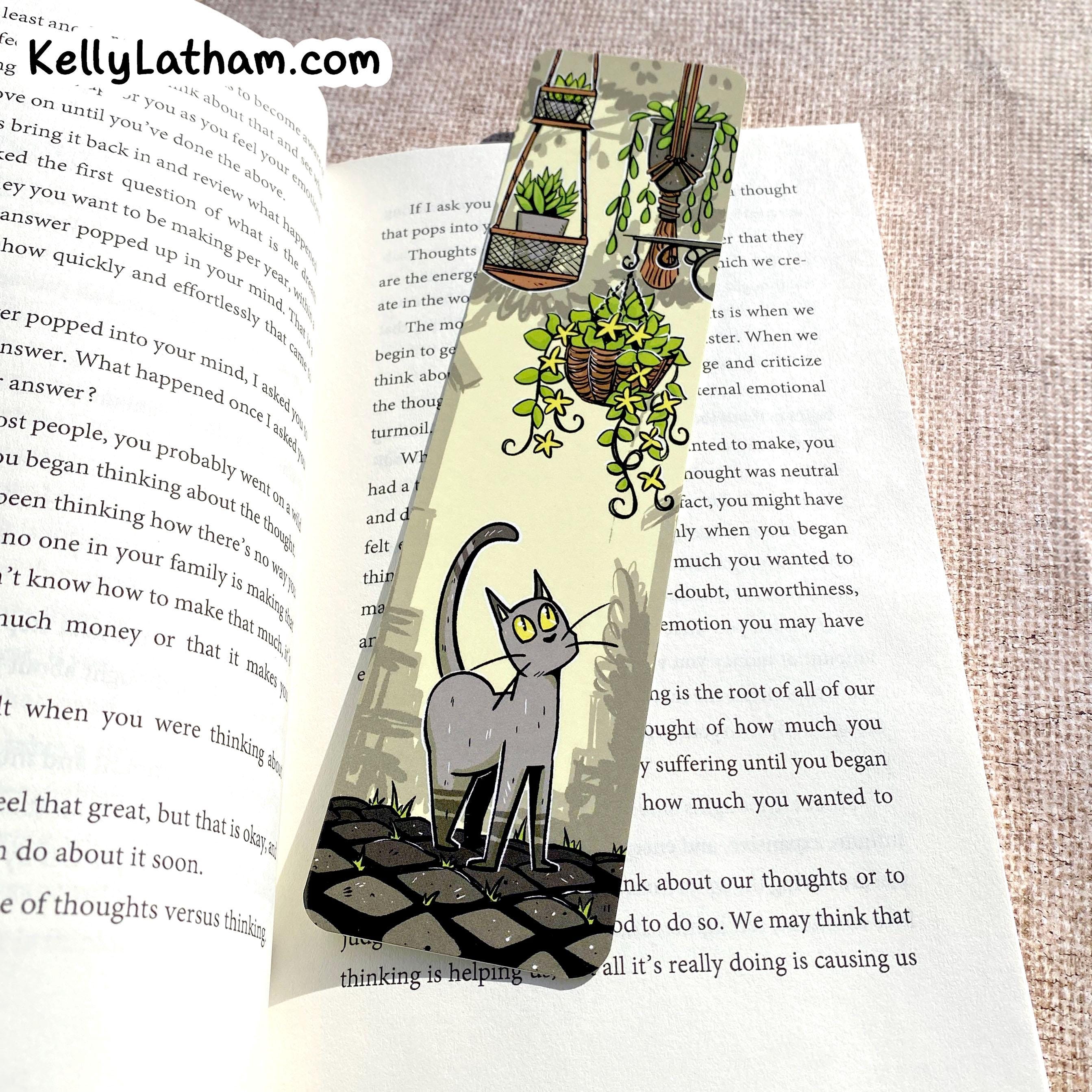 A handmade paper bookmark featuring a black and white cat and potted plants, displayed on an open book over a textured linen background.