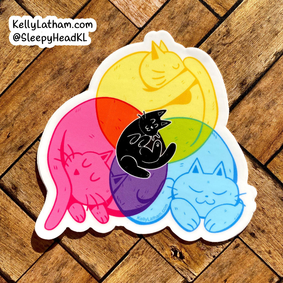 A die-cut sticker featuring four sleeping cats in pink, yellow, blue, and black, layered over overlapping translucent red, orange, green, and purple circles on a wooden floor background.