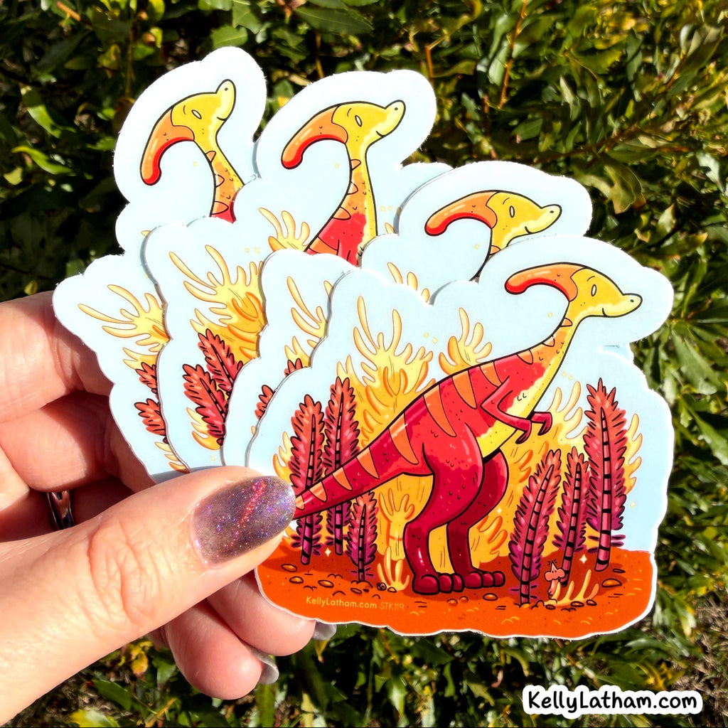 A hand holds a fanned stack of die-cut stickers featuring a red striped dinosaur standing in orange flames with purple plants, against a background of green outdoor foliage.