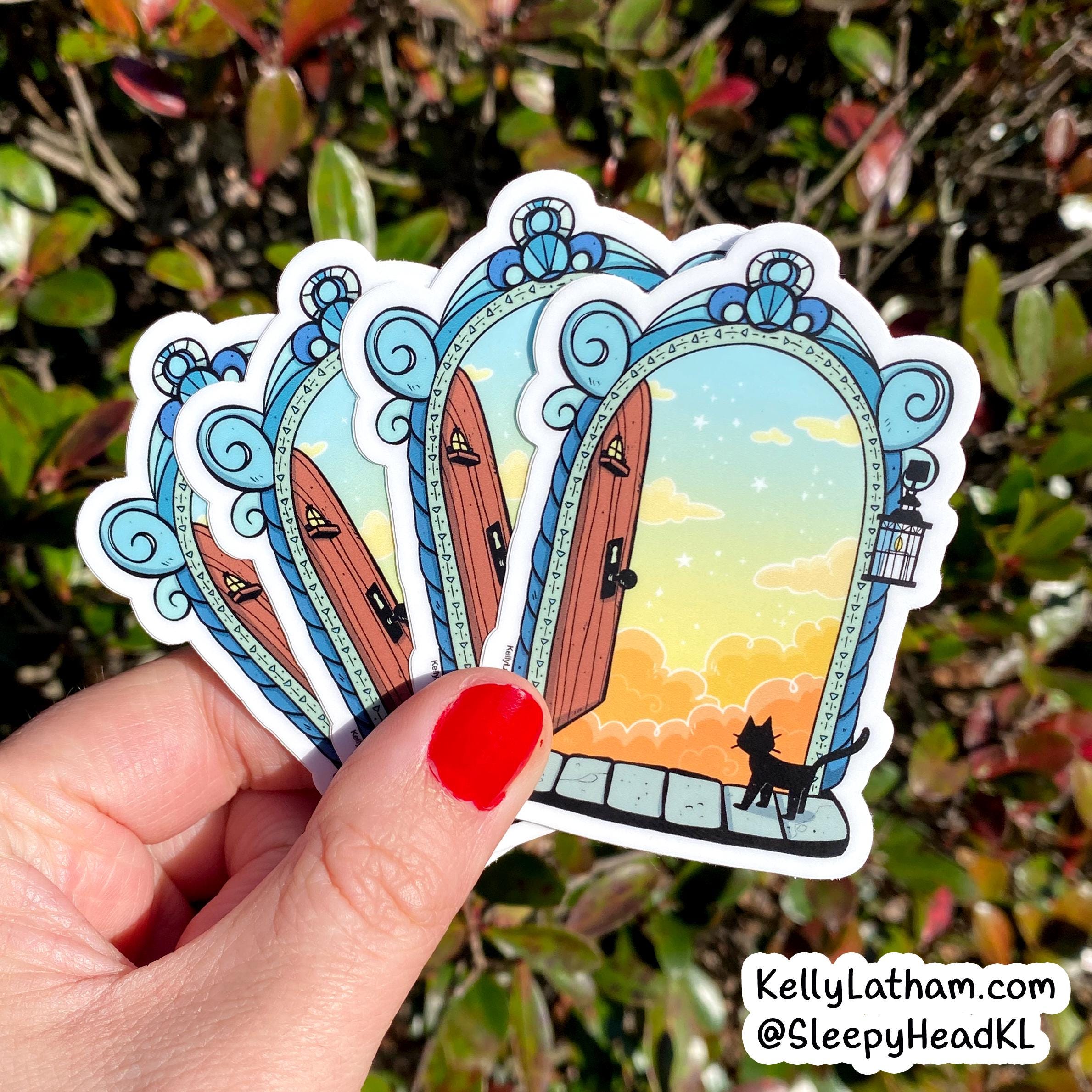 A hand with red nail polish holds four illustrated die-cut stickers featuring ornate blue archways opening to warm sunset skies, with a black cat silhouette at the bottom. The background is blurred green foliage outdoors.